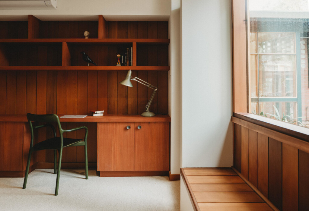 Office space with a wooden desk, built-in shelving, task lamp, and chair beside a window