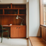 Office space with a wooden desk, built-in shelving, task lamp, and chair beside a window