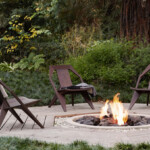Three modern wooden lounge chairs arranged around a bonfire surrounded by greenery