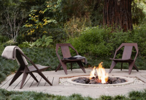 Three modern wooden lounge chairs arranged around a bonfire surrounded by greenery
