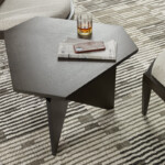 Close-up of a dark wood hexagonal side table holding a glass, newspaper, and phone on a patterned rug between chairs.