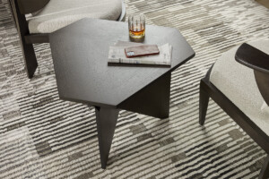 Close-up of a dark wood hexagonal side table holding a glass, newspaper, and phone on a patterned rug between chairs.