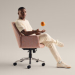 Man sitting in upholstered office chair tossing an orange against neutral background