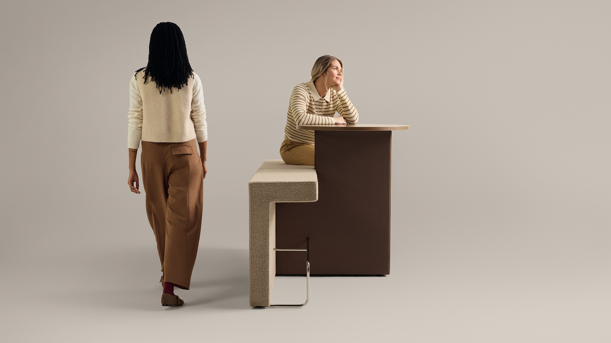 Two women interacting with modern upholstered bench and integrated wood table against neutral background