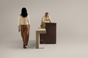 Two women interacting with modern upholstered bench and integrated wood table against neutral background
