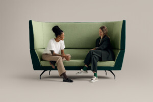 Two people seated on green high-back lounge sofa in conversation against neutral background