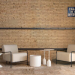 Two beige lounge chairs with black metal arms arranged around small white tables set against a light brick wall.