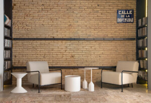 Two beige lounge chairs with black metal arms arranged around small white tables set against a light brick wall.